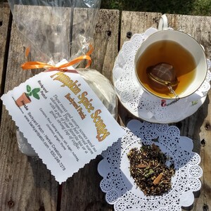 May include: A bag of pumpkin spice scones with a label that reads "Pumpkin Spice Scones, Backyard Patch, Herbal Creations, These sweet and savory scones need no adornment. They are best served warm with a pot of your favorite tea or a good, strong cup of coffee. Contains: flour, baking powder, salt, sugar, nutmeg. www.backyardpatch.com - 630-248-7278". A cup of tea with a tea strainer is on a white doily.
