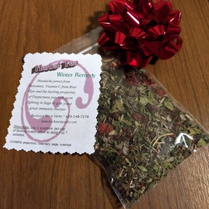 May include: A clear plastic bag filled with dried herbs and berries, tied with a shiny red bow. A white tag with the text "Herbal Tea Winter Remedy" and ingredient information is attached. The tea is a blend of peppermint, rosemary, sage, and rosehips.