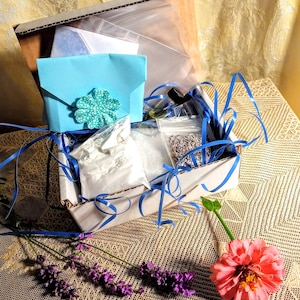 May include: A white cardboard box filled with various items, including a blue envelope with a decorative flower, a bag of white powder, and a bag of lavender buds. The box is surrounded by blue ribbon and is sitting on a brown and white patterned surface.
