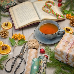 May include: A blue ceramic teacup filled with amber tea on a matching saucer, an open book, two candy canes, metal scissors, a ball of twine, gift-wrapped boxes, pine branches, dried orange slices with star anise, and small red ornaments.