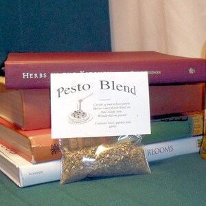 May include: A clear plastic bag filled with dried herbs labeled "Pesto Blend". The bag is sitting on top of a stack of books. The label says "Create a marvelous pesto flavor when fresh basil or time eludes you. Wonderful on pasta! Contains basil, parsley and garlic."