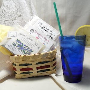 May include: A wicker basket filled with various tea bags, including "Garden Walk Tea", "Sun Gait Tea", and "Ice Tea Brew Calming Spirit". A glass of ice water with a lemon wedge and a green straw is in the foreground.