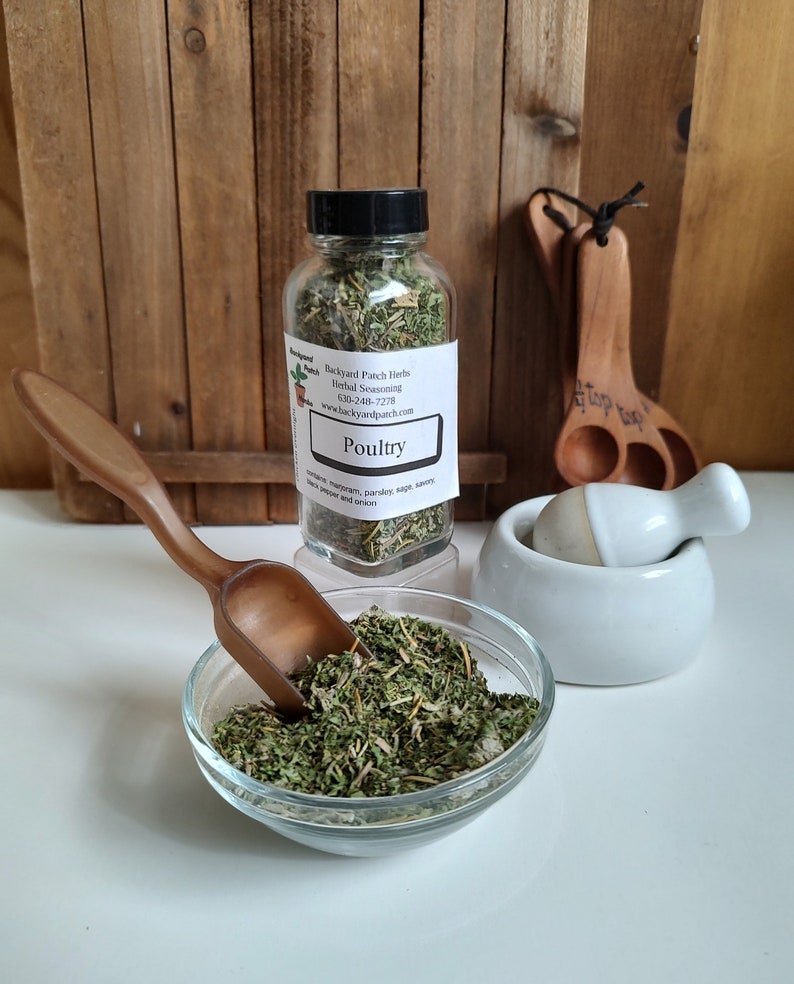 Pu&ograve; includere: Un barattolo di vetro pieno di erbe secche etichettato "Poultry" con i seguenti ingredienti: "coriandolo, maggiorana, prezzemolo, salvia, santoreggia, pepe nero e cipolla". Il barattolo &egrave; su una superficie di legno con un cucchiaio di legno e un mortaio e un pestello bianchi.