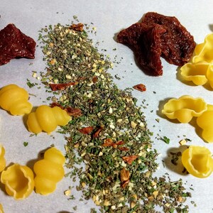 May include: A white surface with a pile of dried herbs and spices, sun-dried tomatoes, and yellow pasta shells.