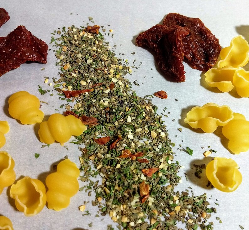 May include: A white surface with a pile of dried herbs and spices, sun-dried tomatoes, and yellow pasta shells.