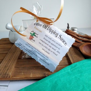 May include: A white label with a gold ribbon tied around it. The label says "Olive Oil Dipping Sauce Tuscan" and has a small image of a potted plant with the text "Backyard Patch Herbs". The label also includes a description of the sauce and its ingredients.