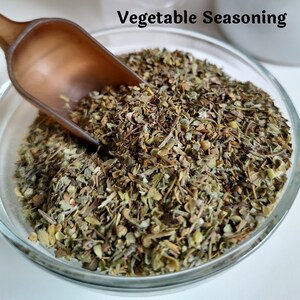 May include: A glass bowl filled with dried herbs and spices, labeled "Vegetable Seasoning". A brown scoop is partially submerged in the bowl.
