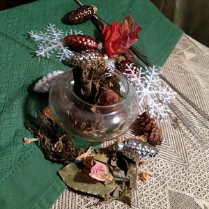 May include: A clear glass bowl filled with dried botanicals, surrounded by decorative elements. Includes pinecone ornaments, white snowflake accents, and a red fabric flower, all arranged on a green and beige patterned surface.