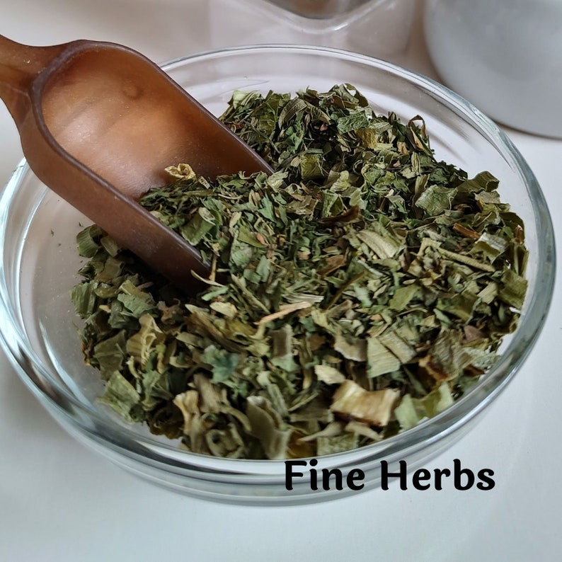 May include: A glass bowl filled with finely chopped green herbs. The herbs are a mix of light and dark green shades. A brown scoop is partially in the bowl. The text "Fine Herbs" is printed below the bowl.