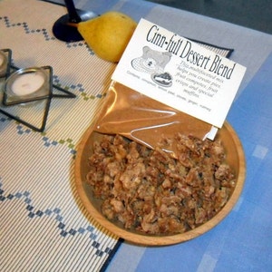 May include: A brown paper bag filled with a cinnamon dessert blend. The bag is labeled "Cinn-full Dessert Blend" and lists the ingredients: cinnamon, lemon peel, cloves, ginger, and nutmeg. The bag is sitting on a wooden bowl filled with brown, crumbly dessert topping.