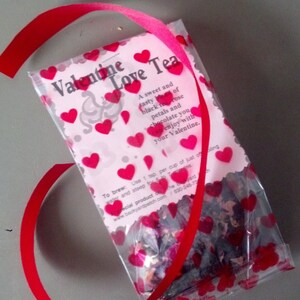 May include: A clear plastic bag filled with loose leaf tea, decorated with red hearts. The bag is tied with a red ribbon and has a label that reads "Valentine Love Tea" with the text "A sweet and tasty blend of black tea, rose petals and chocolate you can enjoy with your Valentine."