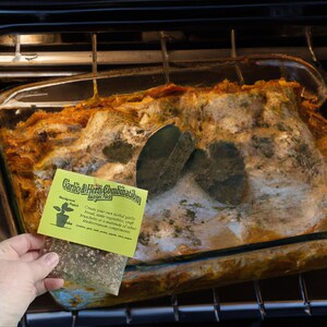 Peut inclure: Un sachet jaune de m&eacute;lange d'assaisonnement &agrave; l'ail et aux herbes s&eacute;ch&eacute;es avec le texte "Garlic & Herb Combination Backyard Patch" et une petite illustration d'une plante. Le sachet est tenu dans une main devant un plat en casserole sale, brun et vert.