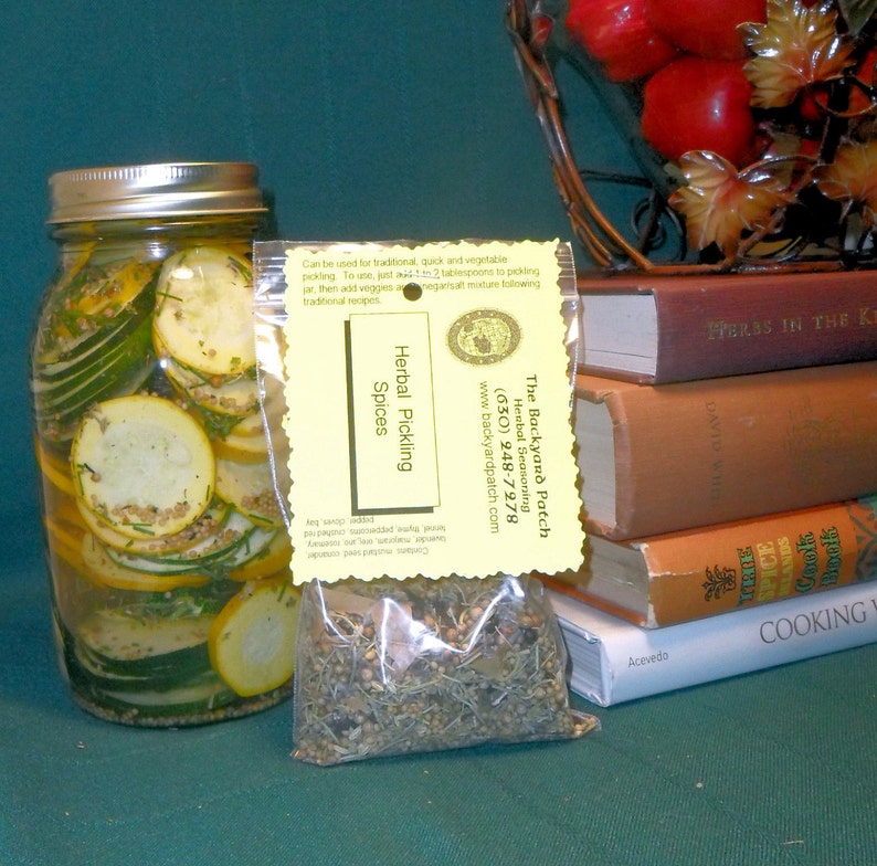 May include: A clear glass jar filled with sliced zucchini pickles. A small yellow bag with a label that reads "The Backyard Patch Herbal Seasoning (630) 248-7978 www.backyardpatch.com Herbal Pickling Spices" is in front of the jar. The bag contains a blend of dried herbs and spices.
