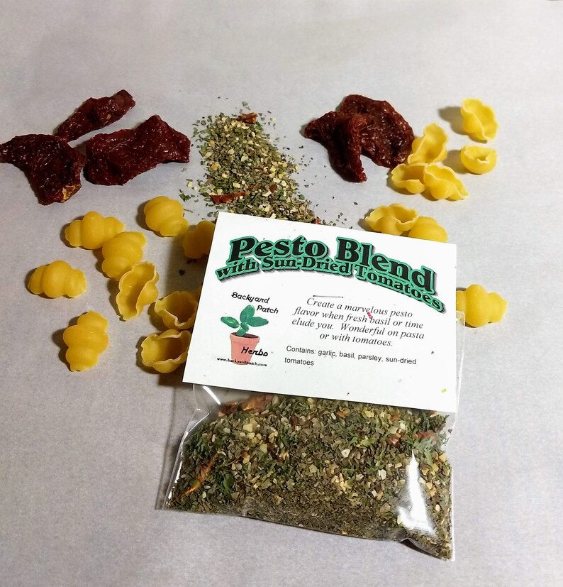 May include: A clear plastic bag filled with a pesto blend with sun-dried tomatoes. The bag has a label that reads "Pesto Blend with Sun-Dried Tomatoes". The label also includes the text "Backyard Patch Herba" and "Create a marvelous pesto flavor when fresh basil or time elude you. Wonderful on pasta or with tomatoes. Contains: garlic, basil, parsley, sun-dried tomatoes."