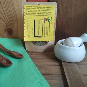 May include: A small brown paper packet of Do-It-All Seasoning from Backyard Patch Herbs. The packet is on a wooden surface with a white ceramic mortar and pestle and a green paper surface. The packet has a yellow label with the text "Do-It-All Seasoning" and "Backyard Patch Herbs" and a website address.