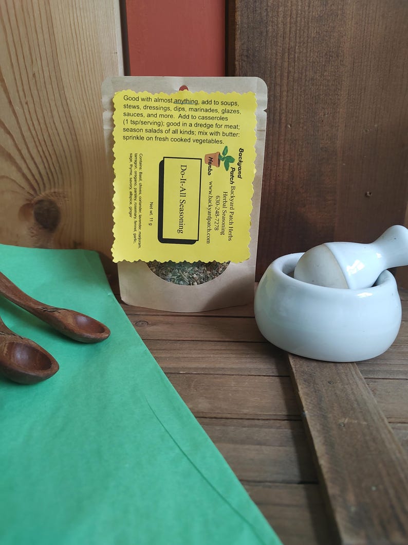 May include: A small brown paper packet of Do-It-All Seasoning from Backyard Patch Herbs. The packet is on a wooden surface with a white ceramic mortar and pestle and a green paper surface. The packet has a yellow label with the text "Do-It-All Seasoning" and "Backyard Patch Herbs" and a website address.