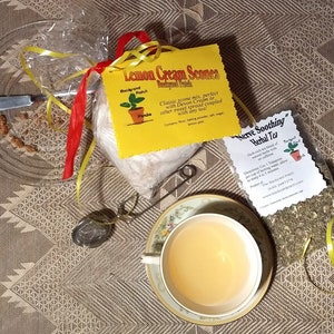 May include: A yellow package with the text "Lemon Cream Scones" and a small bag of dried herbs with the text "Nerve Soothing Herbal Tea". The tea bag is tied with a yellow ribbon. A teacup and saucer are in the background.