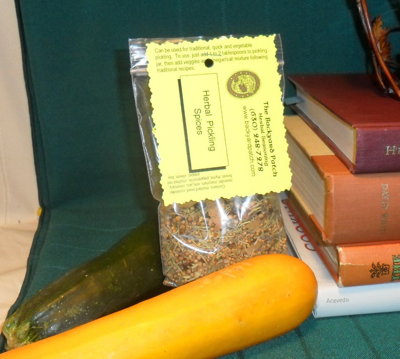 May include: A yellow and green package of Herbal Pickling Spices from The Backyard Patch. The package is sitting on a stack of books. The text on the package reads "Herbal Pickling Spices" and "The Backyard Patch Herbal Seasoning (630) 248-7978 www.backyardpatch.com".