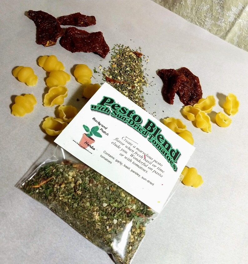 May include: A clear plastic bag filled with a green pesto blend with sun-dried tomatoes. The bag has a white label with the text "Pesto Blend with Sun-Dried Tomatoes" and a description of the ingredients. The label also includes a small illustration of a potted herb plant.