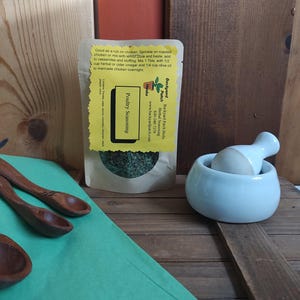May include: A small brown paper bag with a yellow label that reads "Backyard Patch Herbs, Herbal Seasoning, Poultry Seasoning". The bag contains dried herbs and is sitting on a wooden surface with a white mortar and pestle and two wooden spoons.