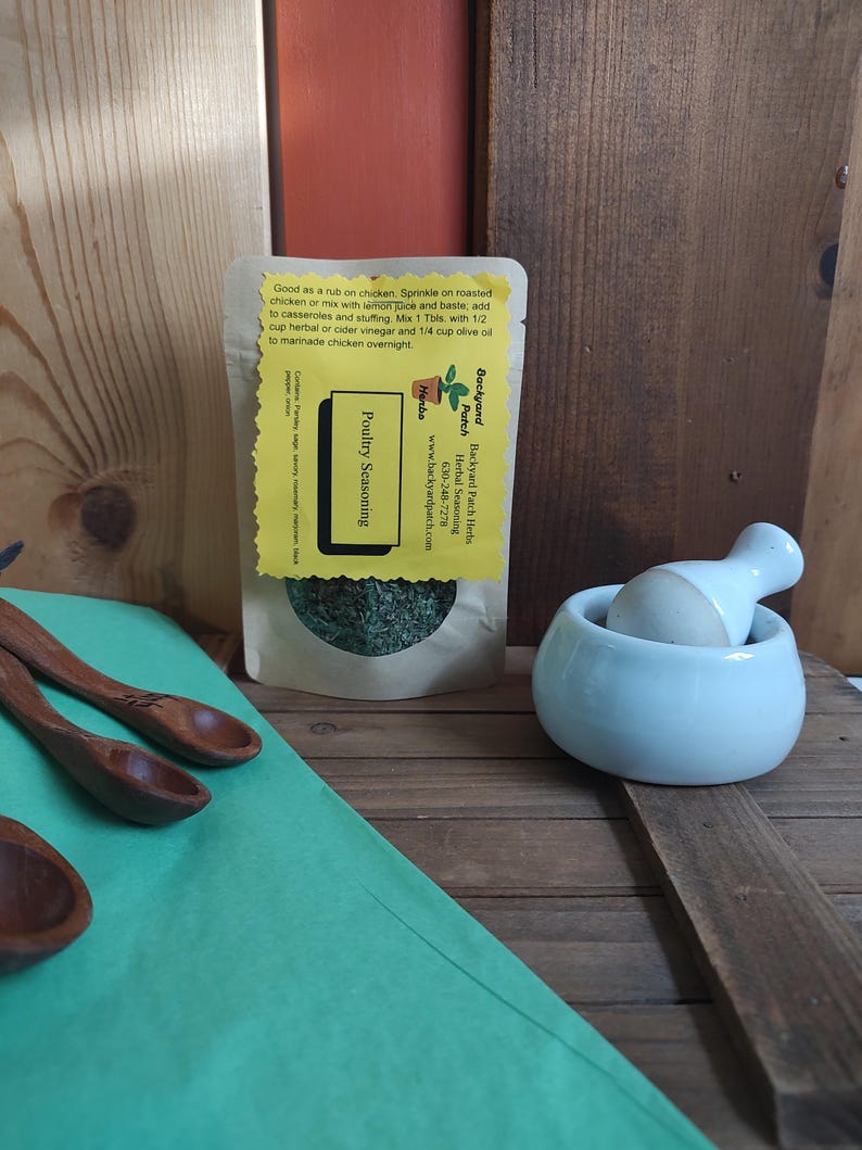 May include: A small brown paper bag with a yellow label that reads "Backyard Patch Herbs, Herbal Seasoning, Poultry Seasoning". The bag contains dried herbs and is sitting on a wooden surface with a white mortar and pestle and two wooden spoons.
