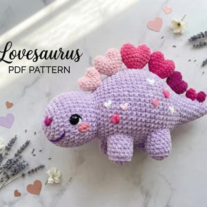 May include: A crocheted purple dinosaur toy with heart-shaped spines and accents. The dinosaur has a smiling face and small white and pink hearts on its body. The text "Lovesaurus PDF PATTERN" is in the upper left corner.