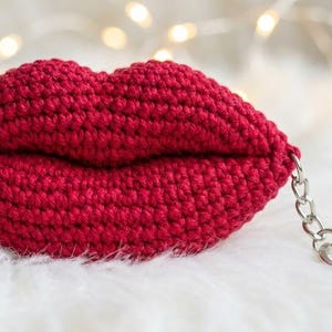 May include: A handmade, red crocheted lip-shaped keychain. The lips are detailed with a defined upper and lower lip, and attached to a silver-colored chain and keyring. The background is a soft, white, fluffy surface.