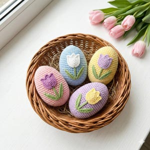 May include: A woven basket holds four crocheted Easter eggs in pastel colors: pink, blue, yellow, and lavender. Each egg features a tulip design in a contrasting color. The basket sits on a white surface, with pink tulips in the background.