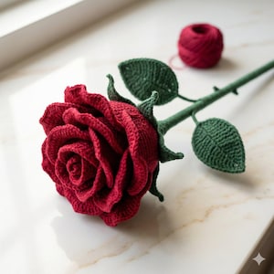 Può includere: Una rosa rossa all'uncinetto fatta a mano, con foglie e stelo verdi. La rosa presenta petali strutturati ed è accompagnata da un gomitolo di filo rosso. Il fiore fatto a mano è un oggetto decorativo.