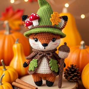 Puede incluir: Figura de zorro de ganchillo con sombrero y capa verdes, sosteniendo un bastón. El zorro está rodeado de calabazas, hojas de otoño y palitos de canela, evocando un tema otoñal.