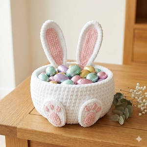 May include: A white crocheted Easter basket in the shape of a bunny, featuring pink-lined ears and paw prints. The basket is filled with foil-wrapped chocolate eggs. A delightful Easter decoration, perfect for treats.