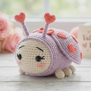 Puede incluir: Una mariquita de ganchillo, de color lavanda con detalles crema y corazones. La imagen incluye una tarjeta de San Valentín, rosas rosas y lana en el fondo.