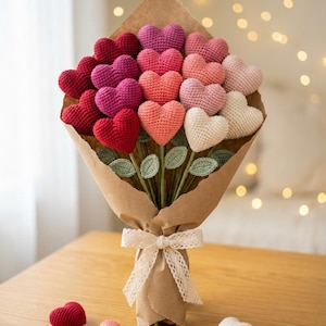 Puede incluir: Un ramo de corazones de ganchillo en tonos rojos, rosas y blancos, envuelto en papel marrón y atado con una cinta color crema. El ramo hecho a mano está sobre una mesa de madera, con algunos corazones sueltos cerca.