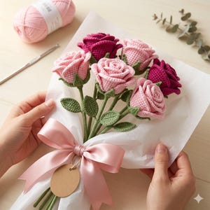 Può includere: Un bouquet di rose all'uncinetto fatte a mano nei toni del rosa e del bordeaux, avvolto in carta bianca e legato con un nastro di raso rosa. Il bouquet include un'etichetta vuota e foglie verdi. Un gomitolo di filato rosa e un uncinetto sono sullo sfondo.