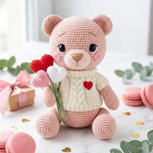 Può includere: Un orsacchiotto rosa all'uncinetto fatto a mano con un maglione color crema con un cuore rosso. L'orso tiene in mano un bouquet di tre cuori all'uncinetto. Lo sfondo include macarons, una confezione regalo e coriandoli a forma di cuore dorato.