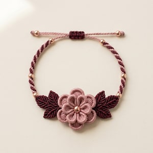 May include: A bracelet featuring a central light pink fabric flower with a metallic bead center, flanked by two dark burgundy fabric leaves. The band is made of twisted burgundy and pink cord, accented with small metallic beads and a sliding knot.