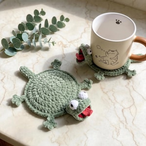 May include: Two handmade sage green crochet frog coasters with white eyes and red tongues. One coaster lies flat, while the other supports a cream-colored ceramic mug featuring a black line drawing of a cat and a small paw print icon.