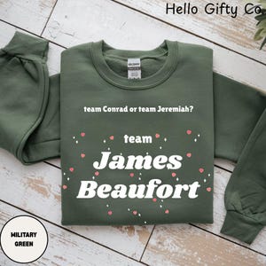 Puede incluir: Una sudadera verde militar con el texto "team Conrad or team Jeremiah?" encima de "team James Beaufort". Pequeños corazones rosas y diamantes blancos decoran el diseño. La sudadera es de cuello redondo.