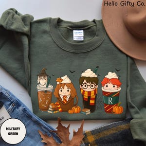 May include: A military green crewneck sweatshirt featuring a graphic print of four cartoon characters in coffee cups with whipped cream toppings, including a wizard, a girl with brown hair, a boy with glasses and a scarf, and a boy with red hair.