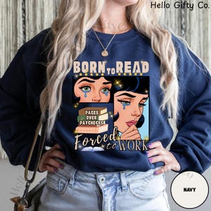 May include: Navy blue sweatshirt with the text "BORN TO READ" above a graphic of two crying women. Below the graphic are the words "PAGES OVER PAYCHECKS" and "FORCED TO WORK". The sweatshirt is a solid navy color.