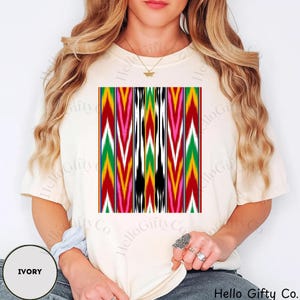 May include: Ivory-colored t-shirt featuring a vibrant, geometric pattern in red, green, yellow, pink, and black. The design is centered on the shirt. The text "Hello Gifty Co." is visible at the bottom right, and "IVORY" is at the bottom left.
