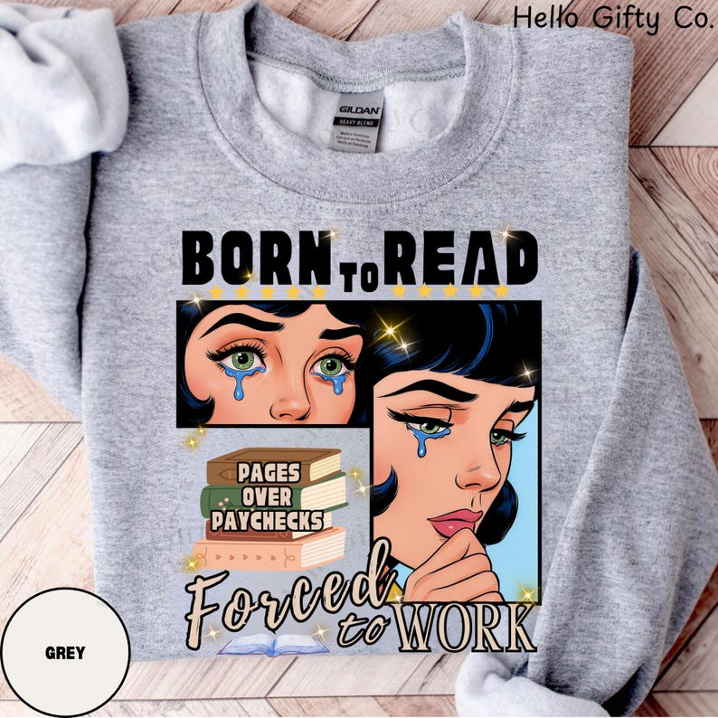 May include: A grey sweatshirt with the text "BORN TO READ" above a graphic of a crying woman. Below the graphic are the words "PAGES OVER PAYCHECKS" and "FORCED TO WORK". The sweatshirt also has the brand name "GILDAN" on the tag.