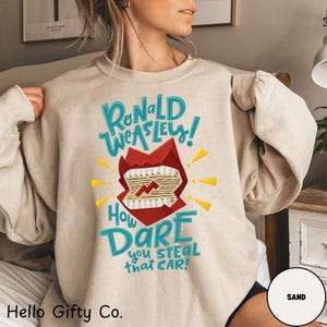 Puede incluir: Sudadera color arena con el texto "Ronald Weasley! How Dare you STEAL that CAR!" en turquesa y blanco. El gráfico incluye una boca roja estilizada con rayos. La marca "Hello Gifty Co." está en la parte inferior.