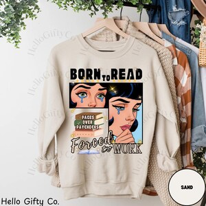 May include: Sand-colored sweatshirt with the text "BORN TO READ" above a graphic of a crying woman. Below the graphic is the text "PAGES OVER PAYCHECKS" and "FORCED TO WORK". The sweatshirt is hanging on a wooden hanger.