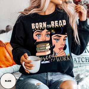 May include: Black sweatshirt with a graphic that reads "BORN TO READ" above a comic-style illustration of a crying woman. The graphic also includes the text "PAGES OVER PAYCHECKS" and "FORCED TO WORK". A white mug is held in the hand.