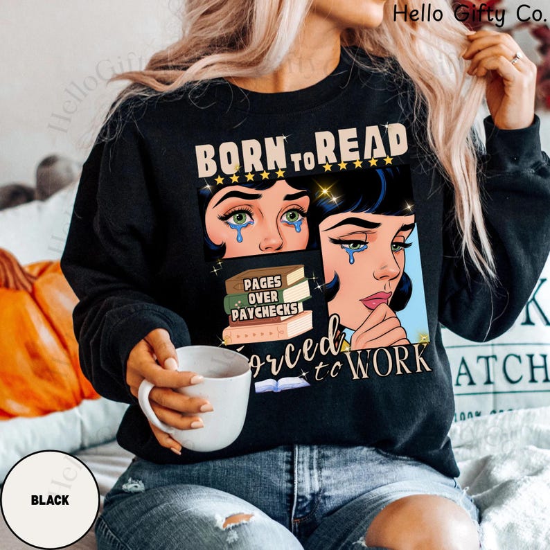 May include: Black sweatshirt with a graphic that reads "BORN TO READ" above a comic-style illustration of a crying woman. The graphic also includes the text "PAGES OVER PAYCHECKS" and "FORCED TO WORK". A white mug is held in the hand.