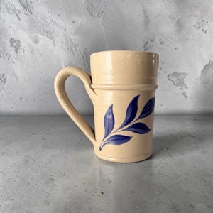 May include: A beige ceramic mug with a speckled finish and a single curved handle. The mug features a hand-painted dark blue leaf motif on the front, with a raised horizontal band encircling the upper portion of the cylindrical body.
