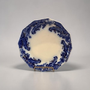 Puede incluir: Un plato octogonal vintage con un centro color crema y un patrón de olas azules alrededor del borde. El diseño azul está acentuado con detalles dorados. El plato se exhibe sobre un soporte de metal dorado.