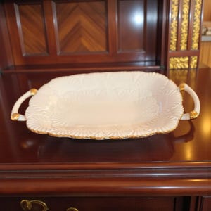 Puede incluir: Bandeja rectangular color crema con borde festoneado y dos asas. La bandeja presenta un relieve floral y ribete dorado. Una pieza decorativa para servir alimentos o exhibir objetos.