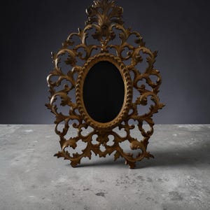 May include: Ornate gold-colored picture frame with an oval opening and intricate scrollwork design. The frame stands upright on a concrete surface, showcasing its detailed craftsmanship and vintage aesthetic. The dark background enhances the frame's elegant appearance.